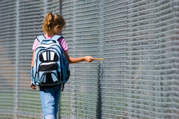 Little girl walking to school by school fence on beautiful sunny day wth back pack and pencil